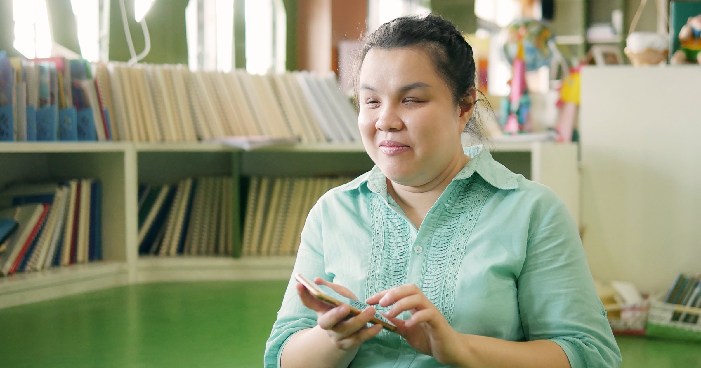 A visually impaired woman smiles as she uses her phone in a library, with shelves of books behind her.