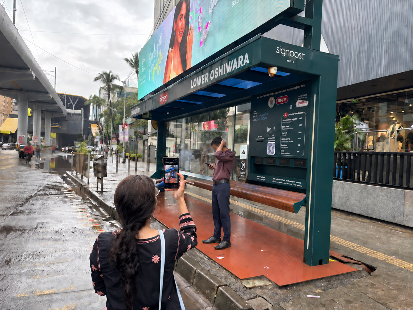 A woman holds up her phone running Audivision to read the Lower Oshiwara bus stop sign on a rainy Mumbai street, while people wait at the shelter.